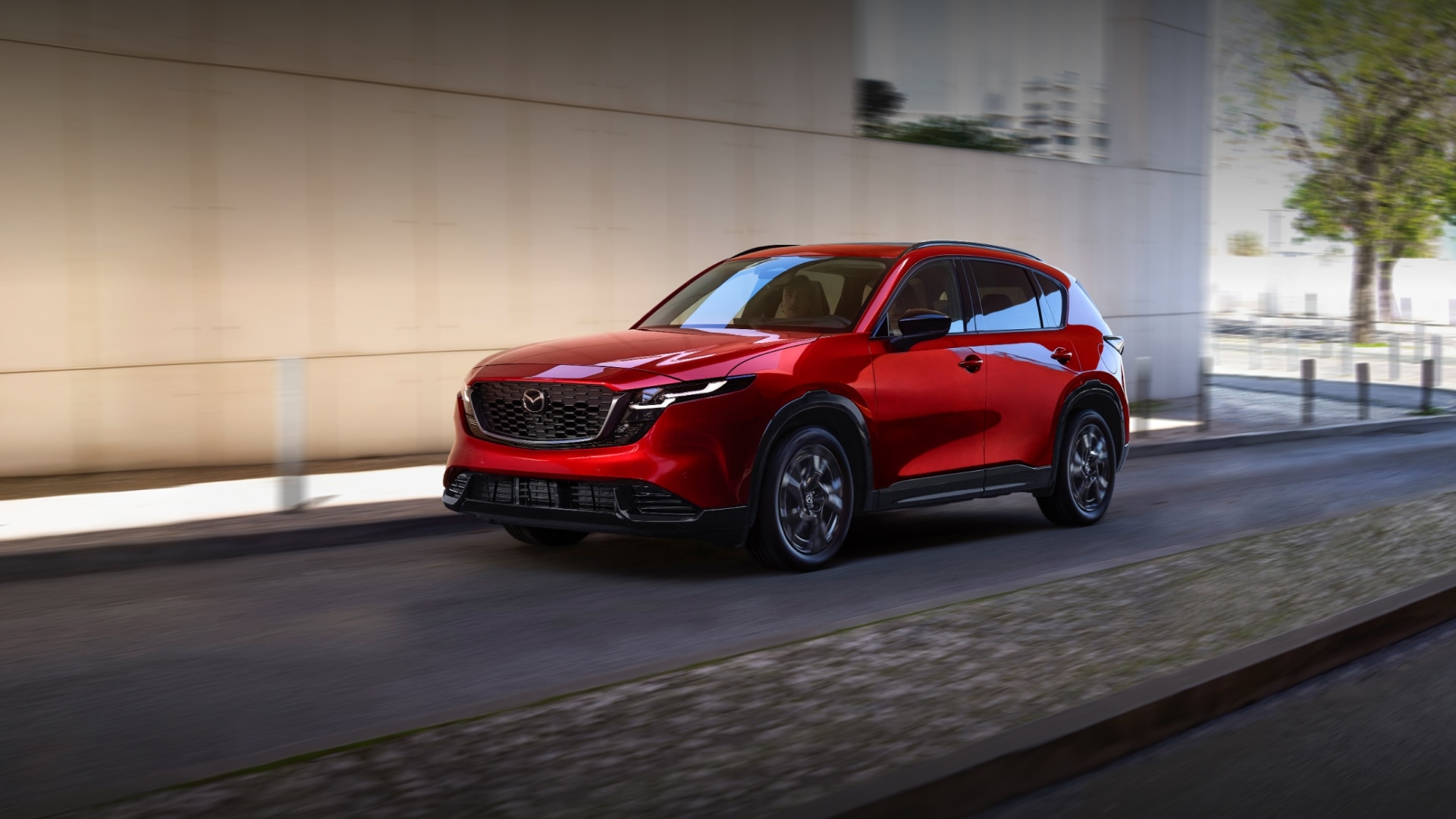 Red Mazda CX-5 SUV driving down a city street alongside a modern building.