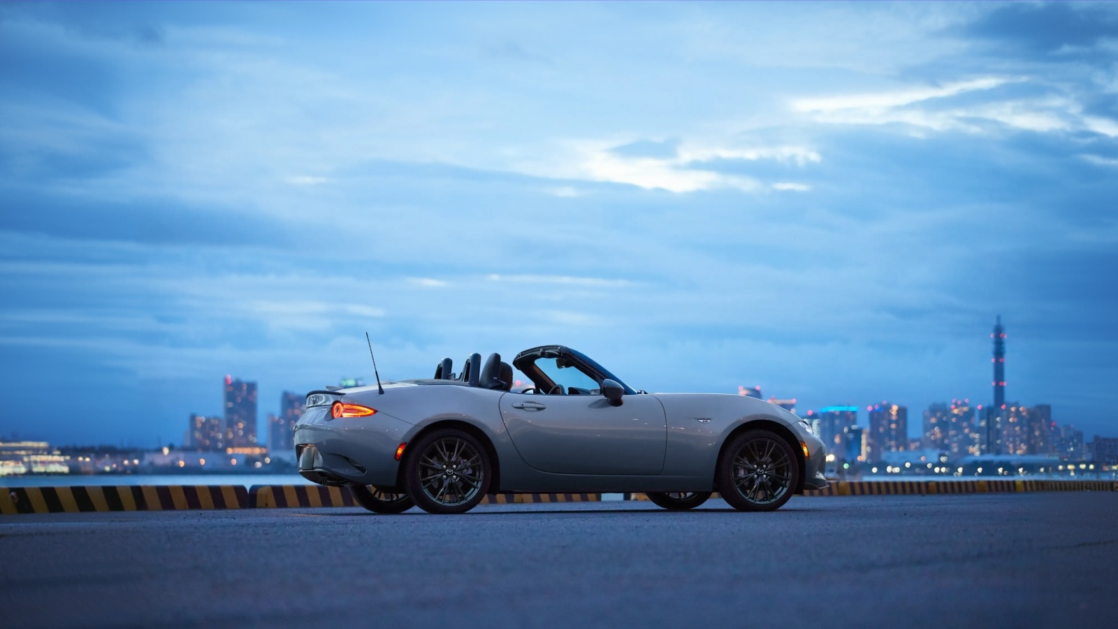 Grey Mazda MX-5 Miata convertible parked on a waterfront with a city skyline at twilight.