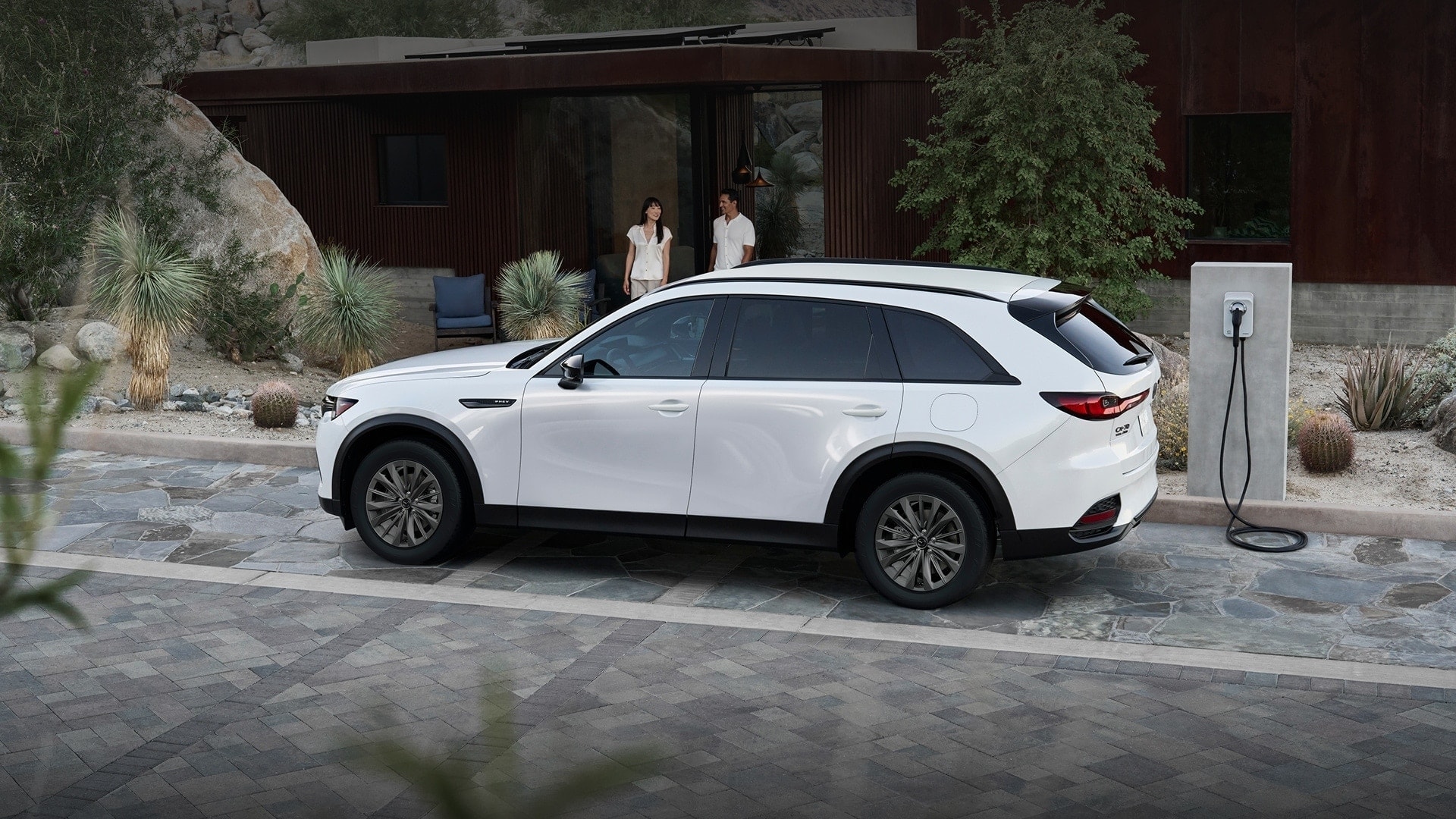 White Mazda CX-70 PHEV SUV parked on a stone driveway next to an EV charging station.